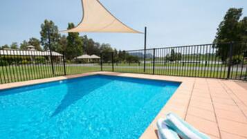 A swimming pool with a tan shade sail, a black metal fence, and a tiled deck.