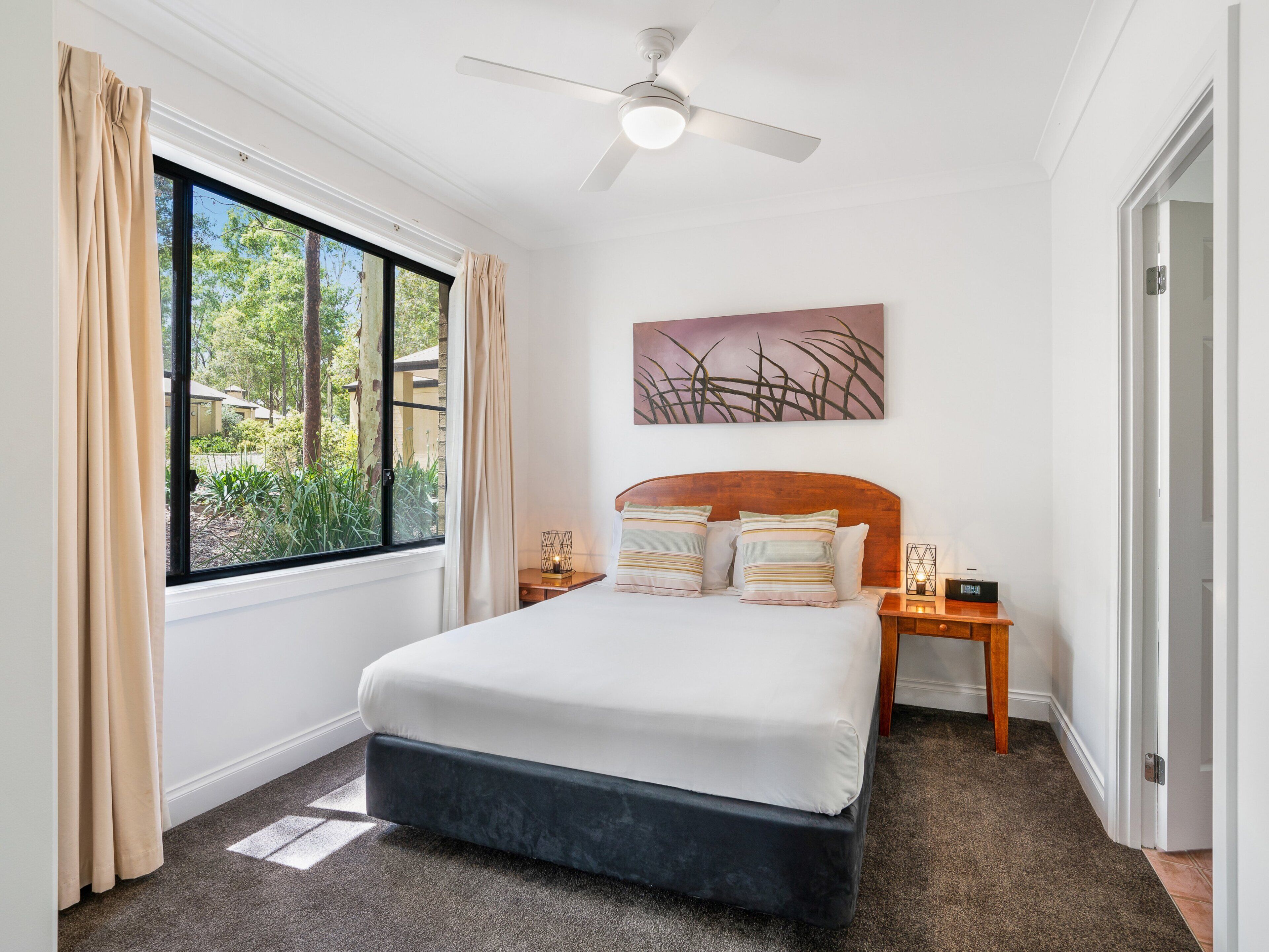 A bedroom with a bed, a ceiling fan, a window with curtains, a nightstand, and a wall-mounted artwork.