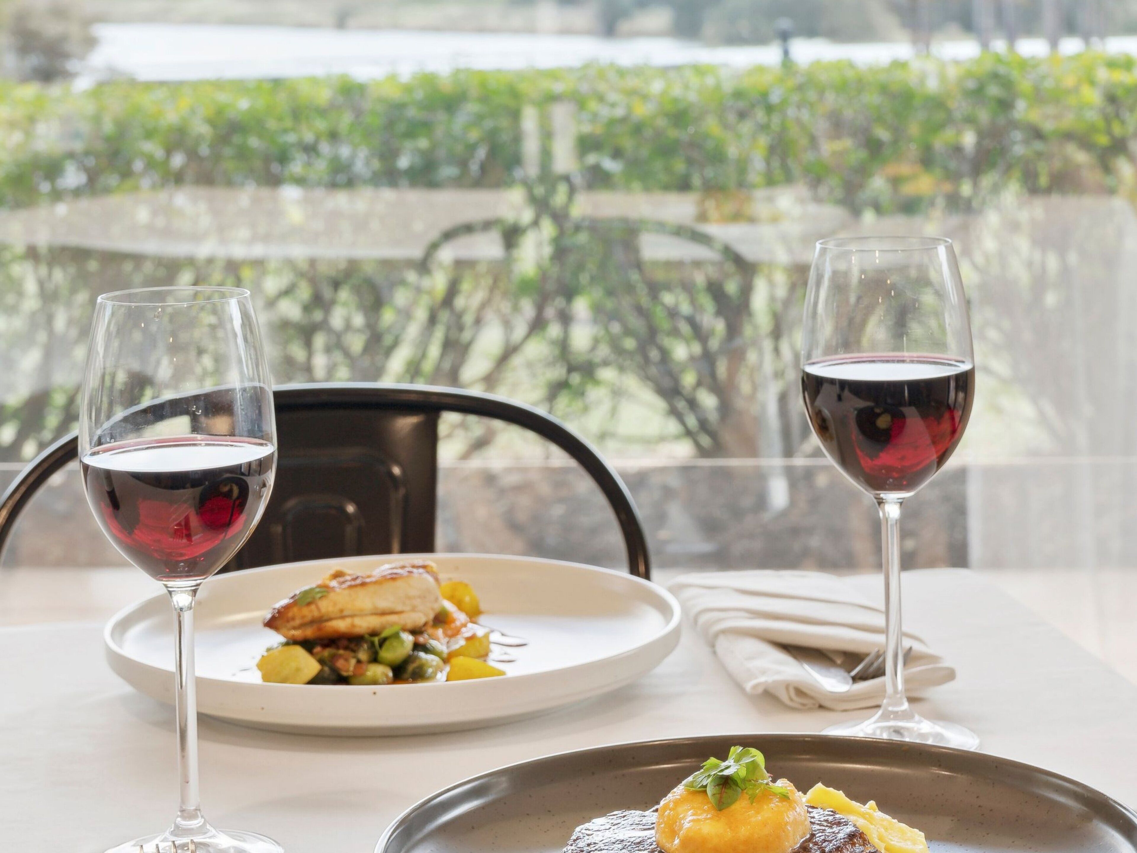 A table with two wine glasses and two plates of food, a chair, and a view of greenery through a window.