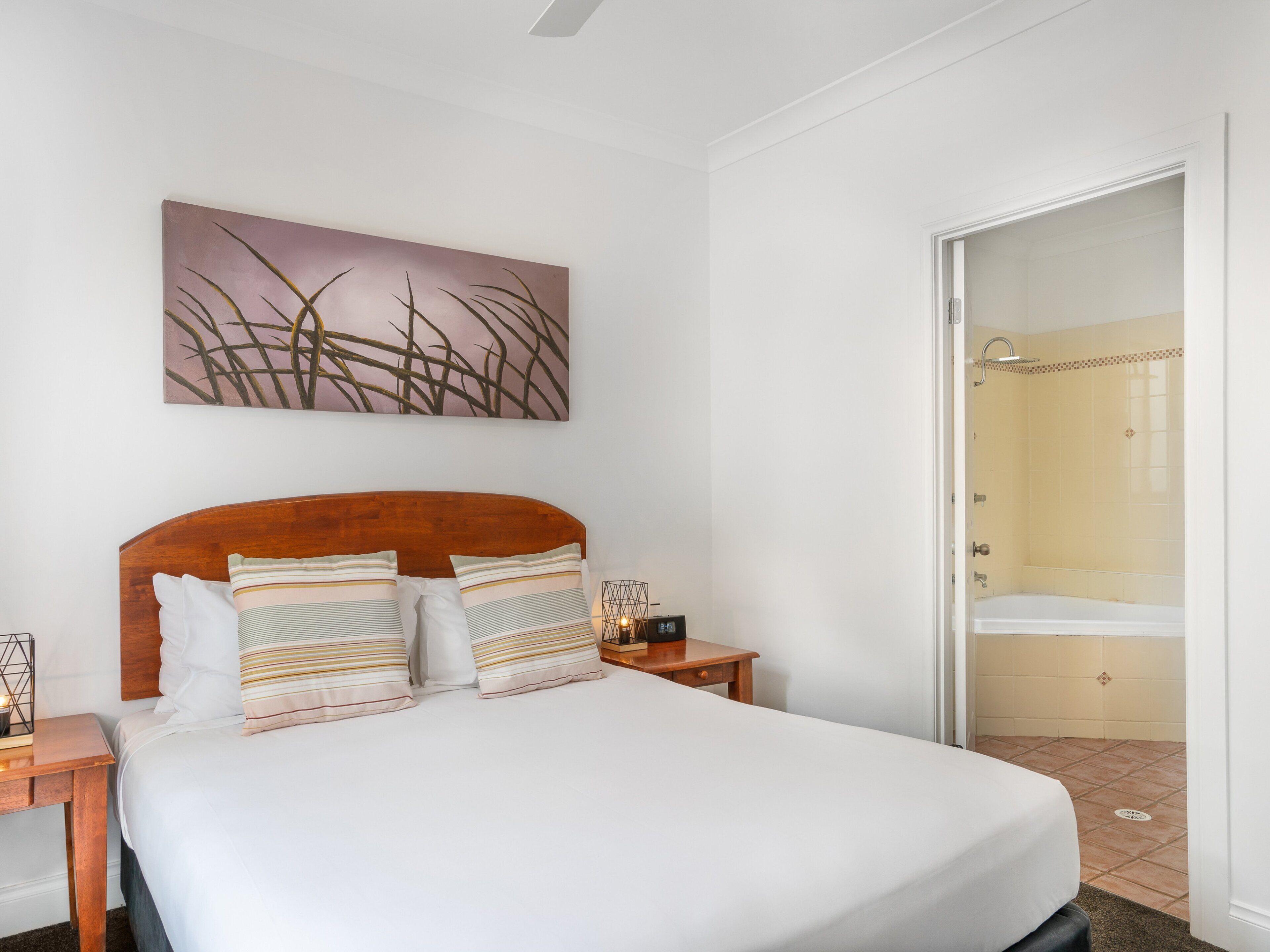 A neatly arranged hotel room with a large bed, a bedside table with a lamp, a wall-mounted artwork, and a bathroom visible through an open door.