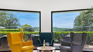 Una habitación con dos ventanas amplias, una mesa redonda de madera con una botella y copas, y dos sillones, uno amarillo y otro gris.