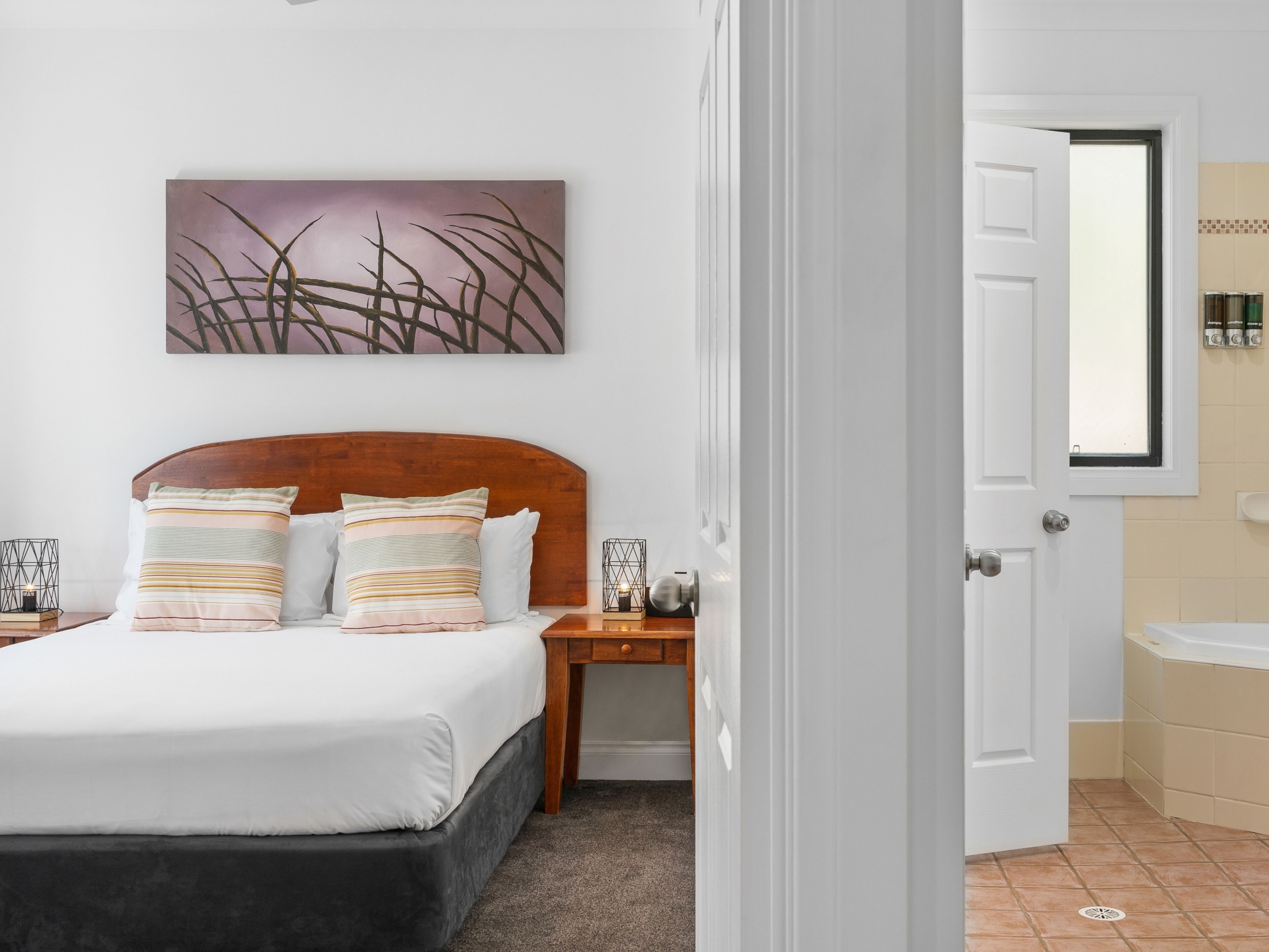 A bedroom with a bed, bedside tables, a nightstand, a wall-mounted artwork, and a bathroom visible through an open door.