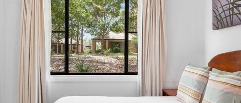 Un dormitorio con un ventanal grande, una cama con almohadas a rayas y vistas a un jardín y árboles.