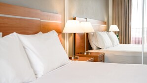 Habitación de hotel con dos camas, cada una con sábanas y almohadas blancas. Hay dos mesitas de noche con lámparas. La habitación tiene un cabecero de madera y un ventanal con cortinas.