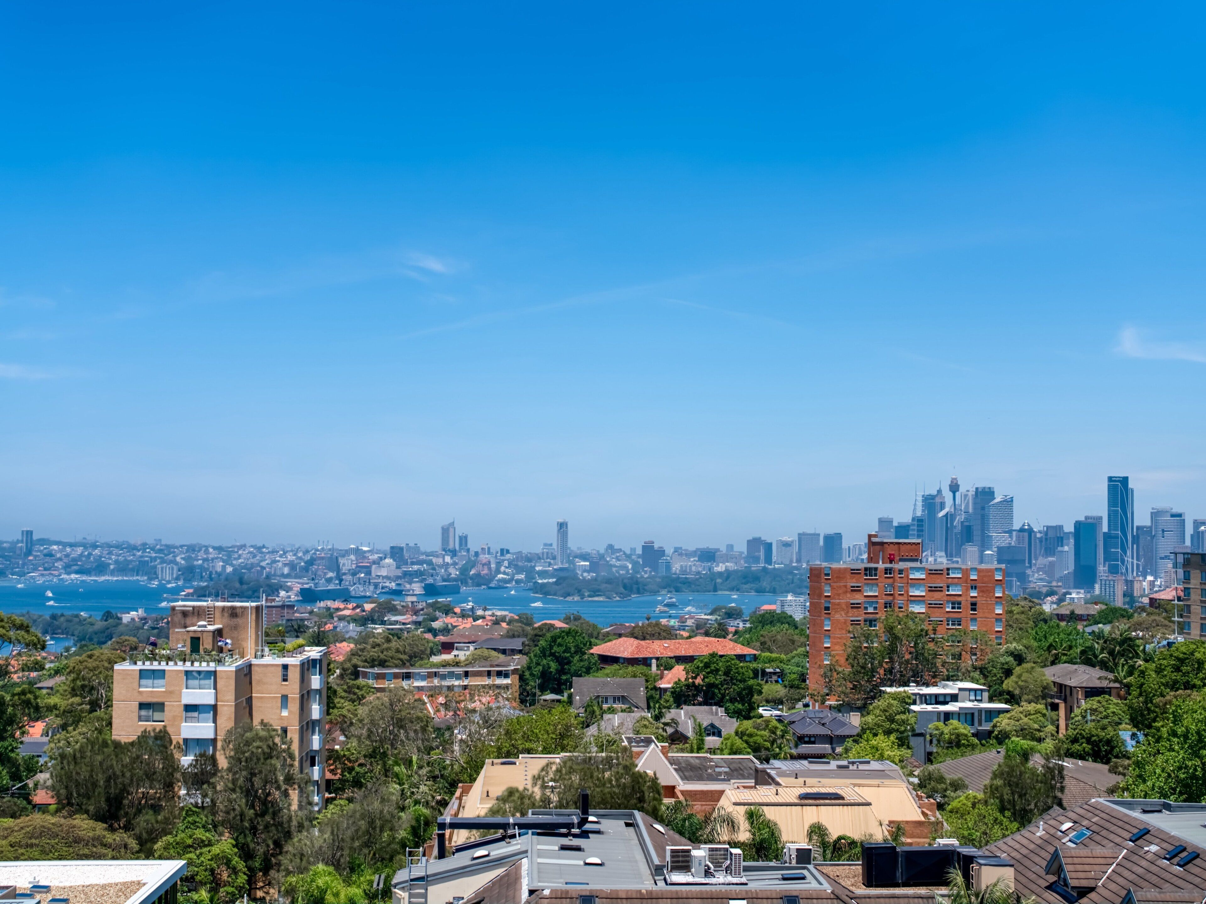 A cityscape with a mix of residential and commercial buildings, a clear blue sky, and a body of water in the distance.