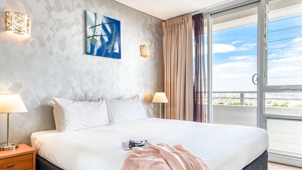 Una habitación de hotel con una cama grande, dos lámparas de noche, un cuadro en la pared y vistas a través de una puerta corrediza de vidrio.