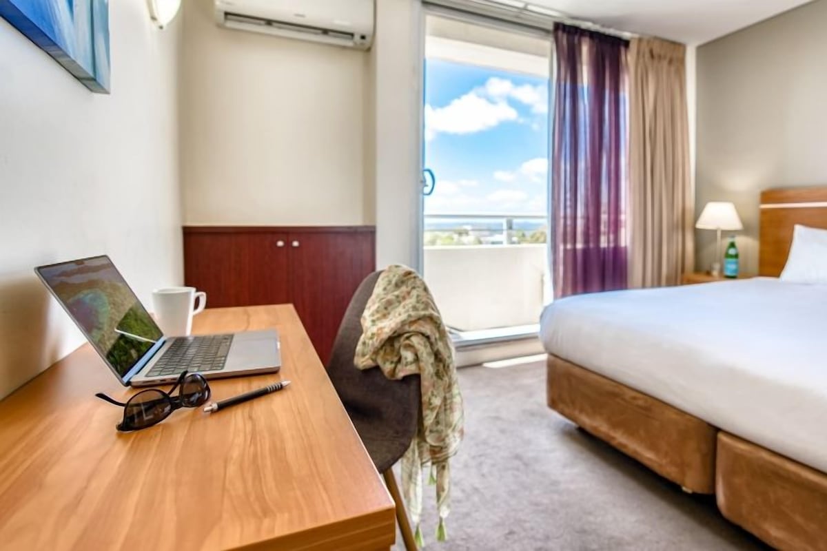 A hotel room with a bed, a desk with a laptop, a chair, a window with curtains, and a view of the sky.