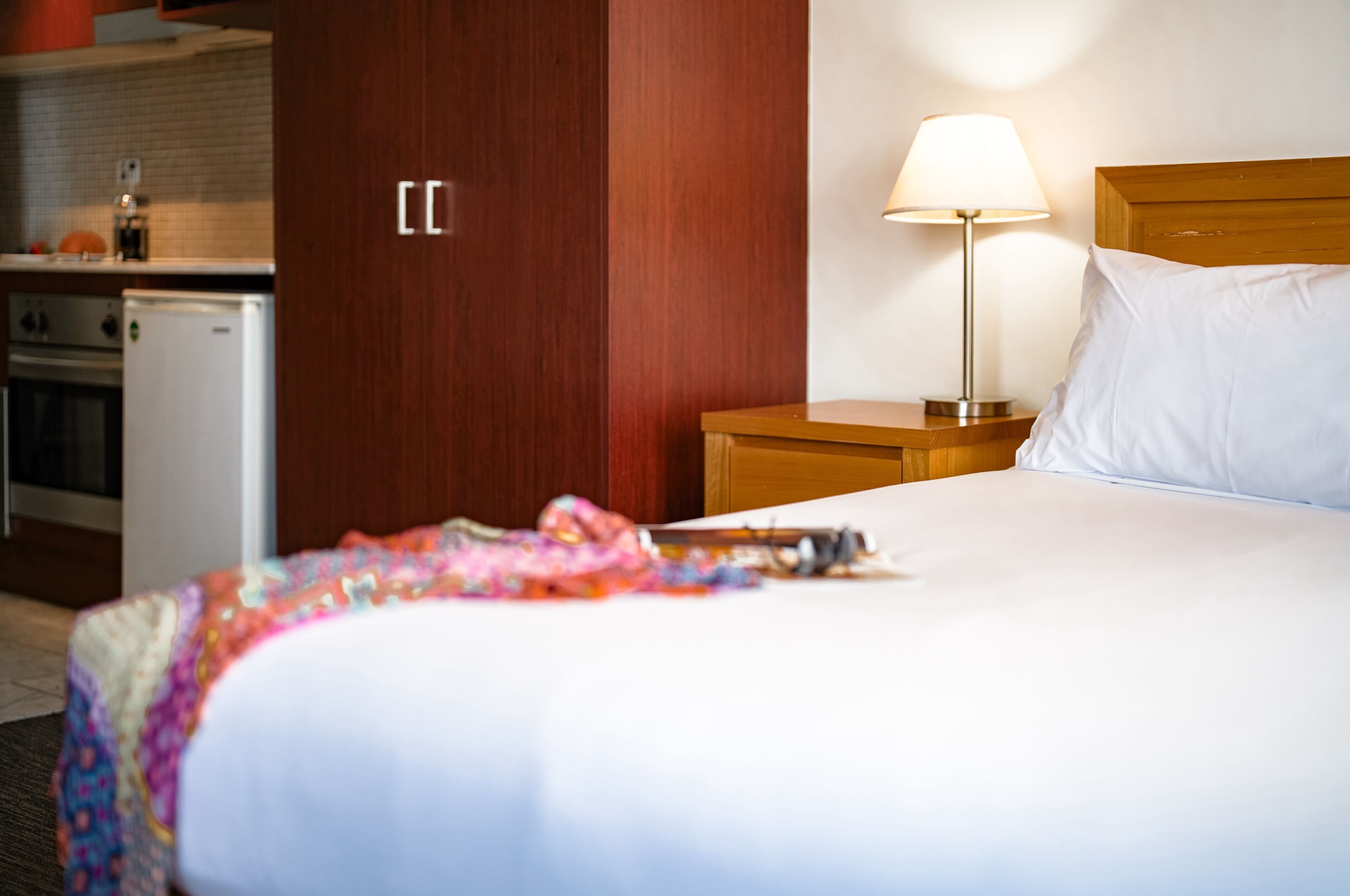 A hotel room with a bed, a nightstand with a lamp, a wooden headboard, a small kitchenette with a refrigerator and stove, and a door with a handle.