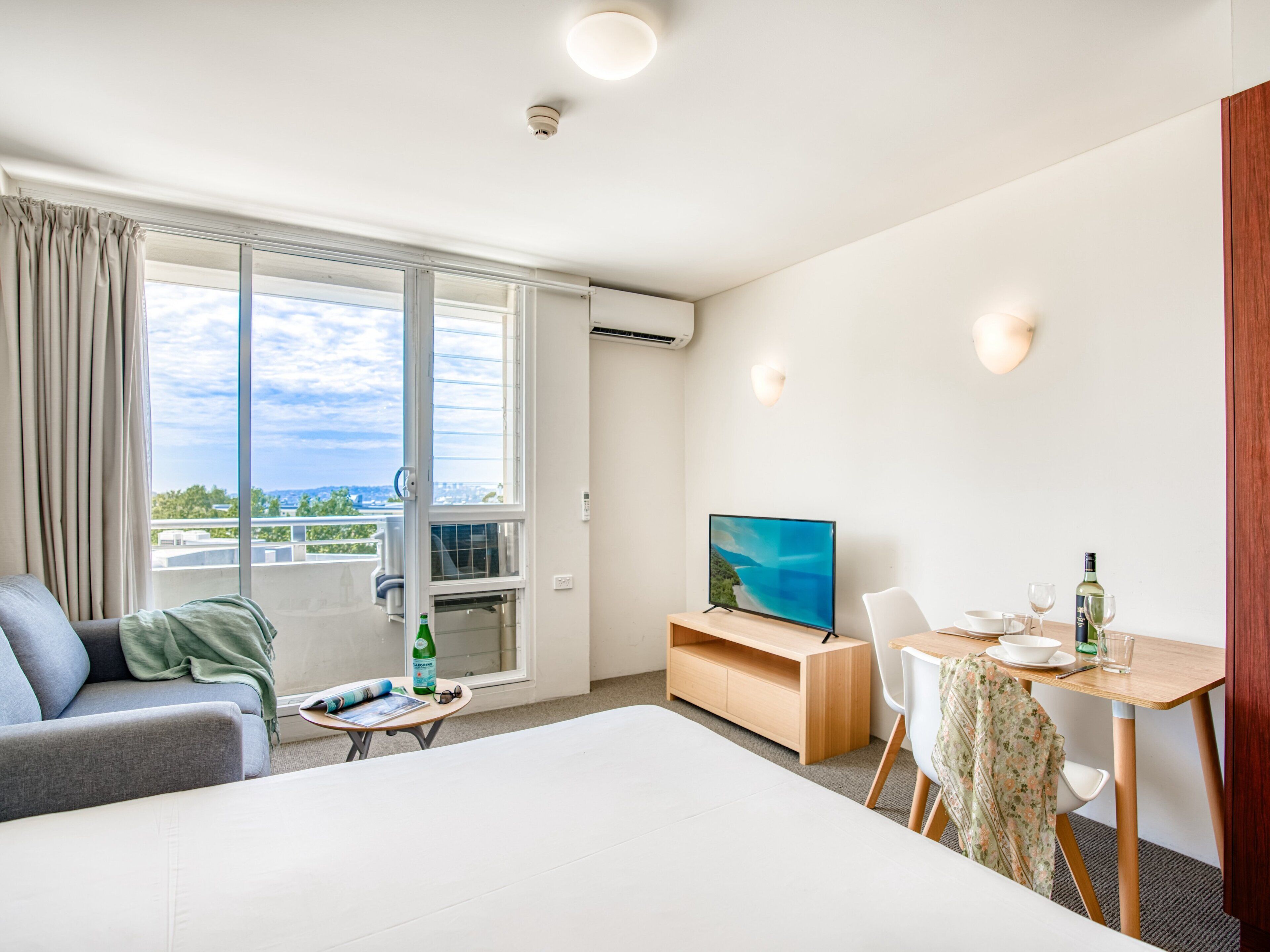A modern hotel room with a large bed, a sofa, a dining table with chairs, a TV, and a balcony with a view of the city.