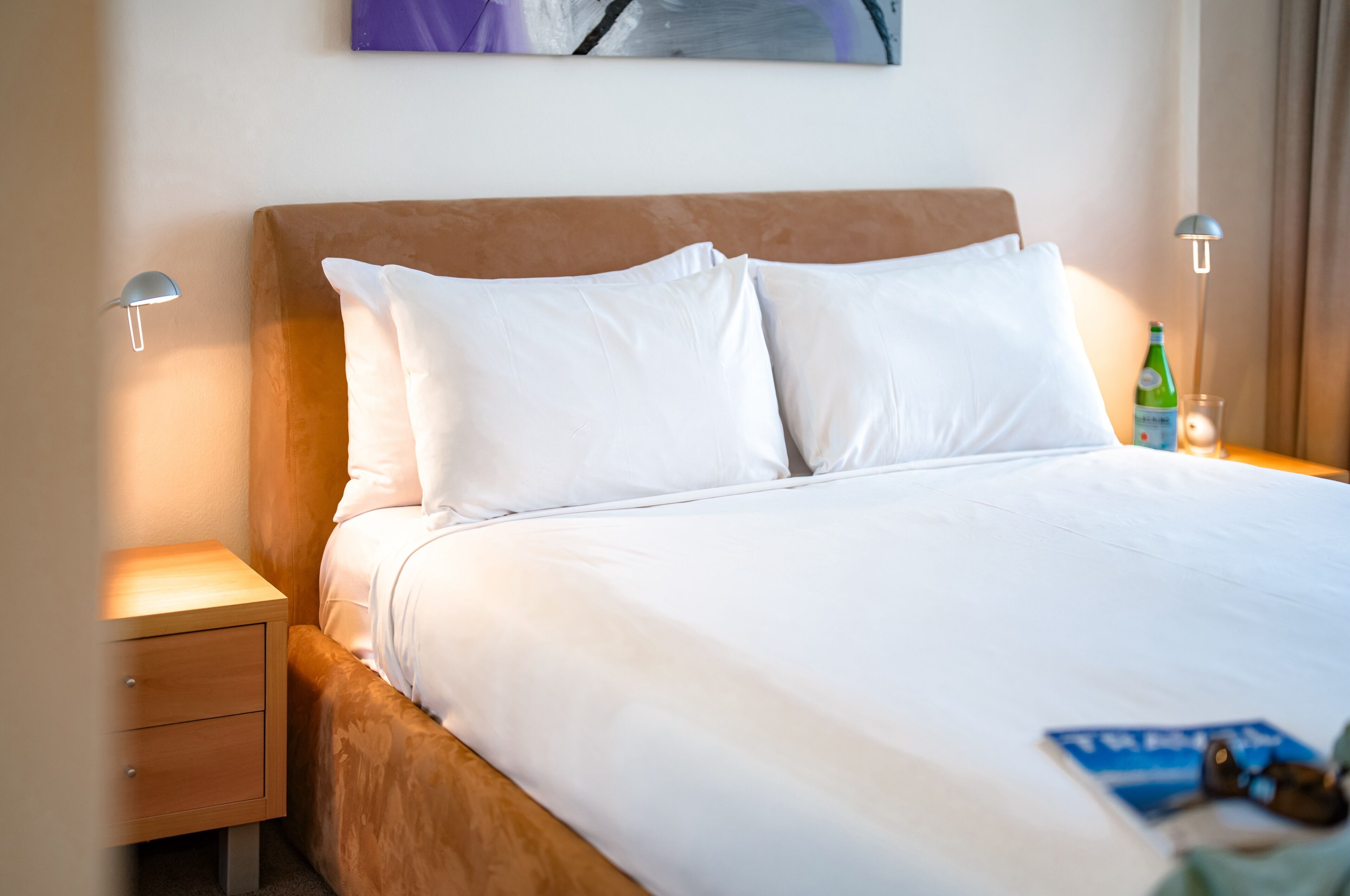 Un lit bien fait, avec du linge de lit blanc, une tête de lit en bois et une petite table de chevet sur laquelle se trouve une lampe.