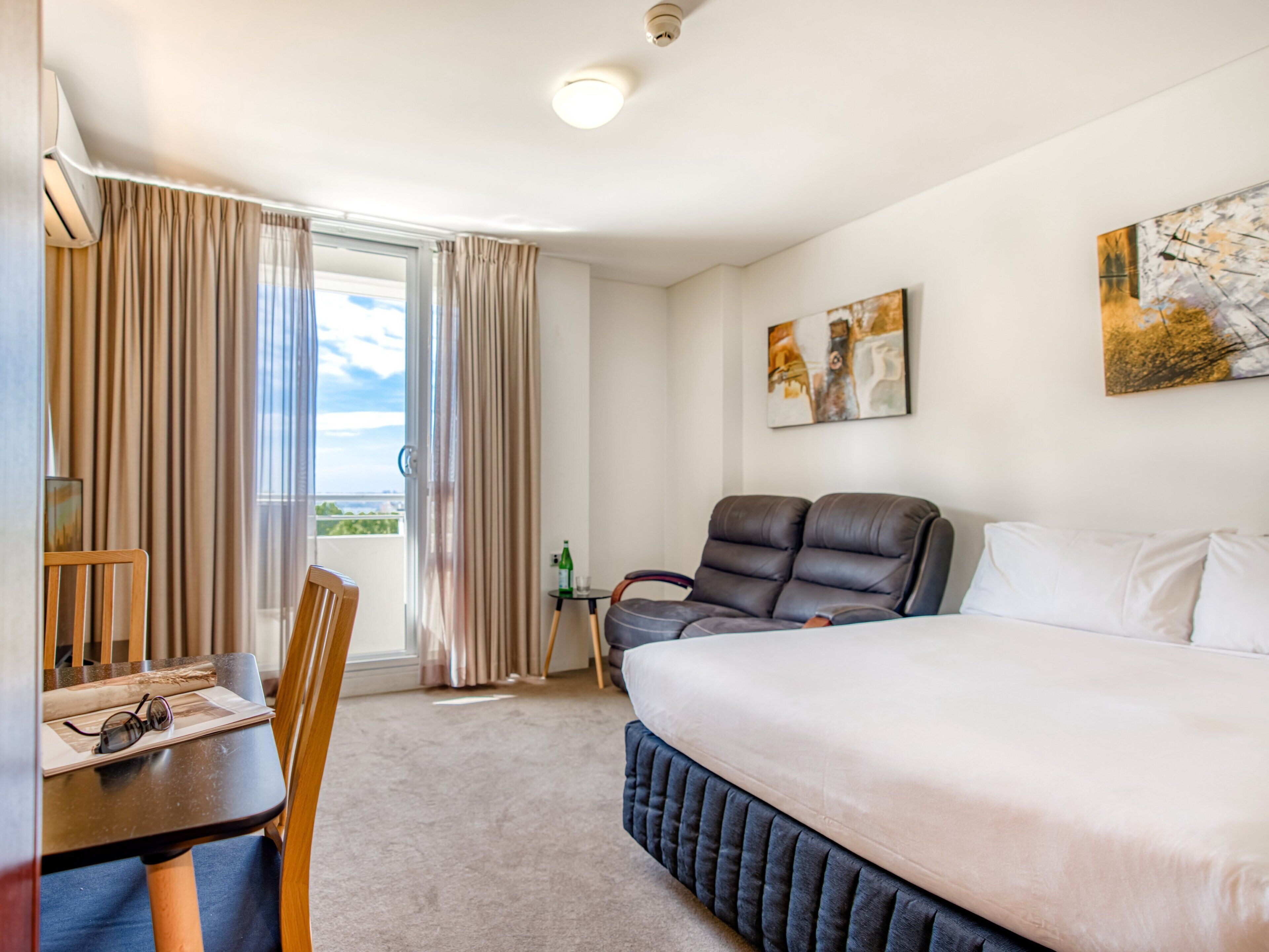 A hotel room with a bed, a desk, a chair, a massage chair, and a view of the outdoors.