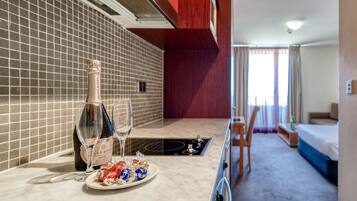 A hotel room with a kitchen area, a dining table with a bottle of champagne and glasses, a plate of assorted chocolates, and a bed with a blue bedspread.