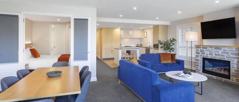 A modern living room with a fireplace, blue sofas, a dining area, and a kitchen in the background.