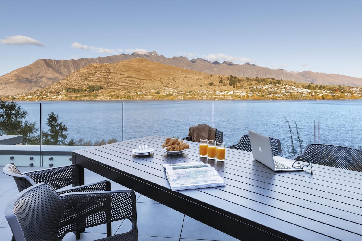 Una mesa de comedor con una computadora portátil, vasos y alimentos para el desayuno, con vista a un lago y montañas.