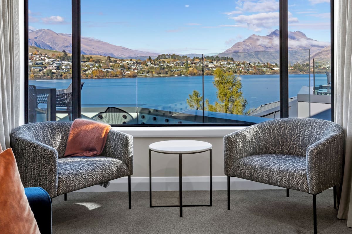 Una habitación moderna con dos sillones estampados, una mesita redonda y un ventanal con vistas a un lago y montañas.