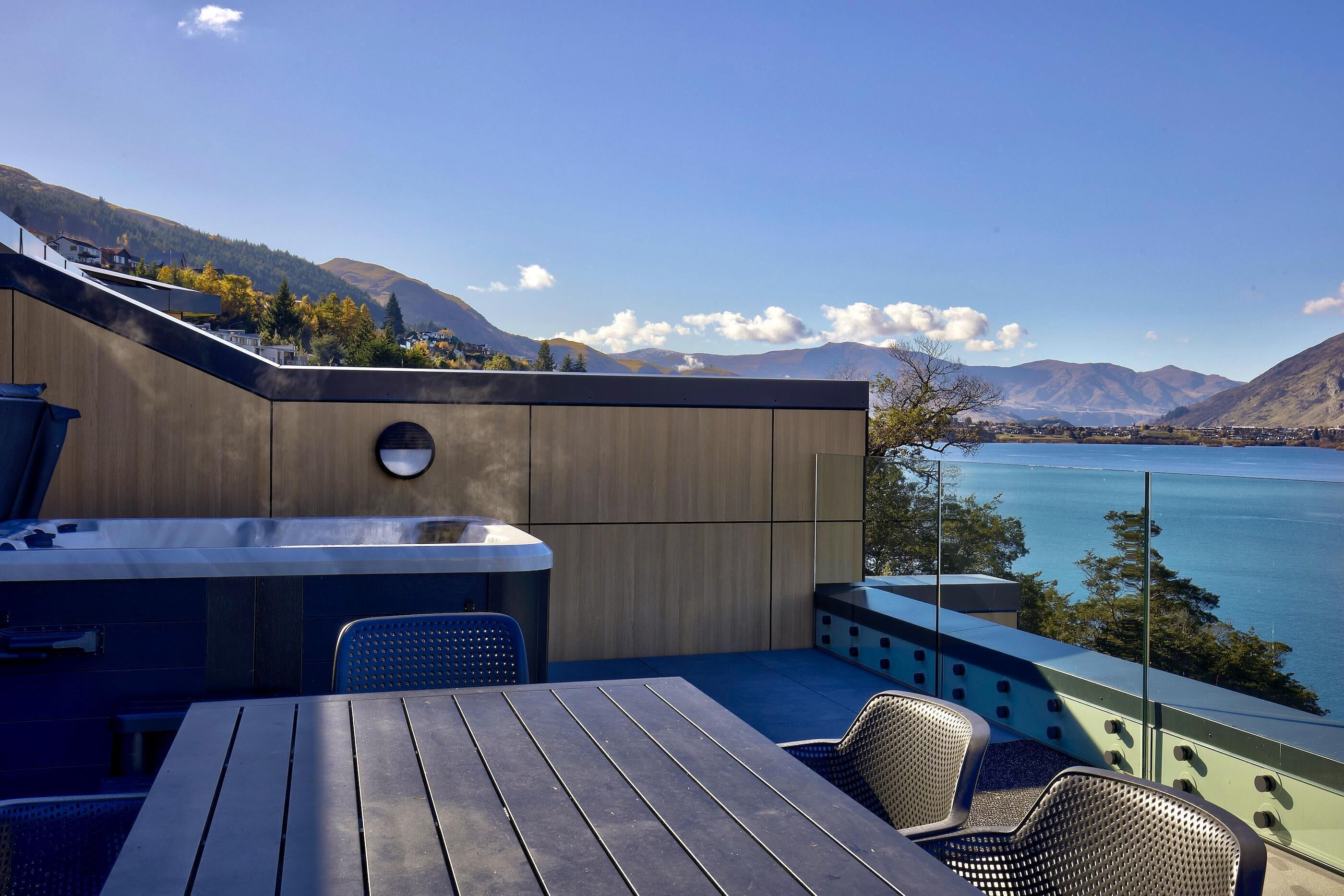 A modern outdoor seating area with a wooden table and chairs, overlooking a lake and mountains.
