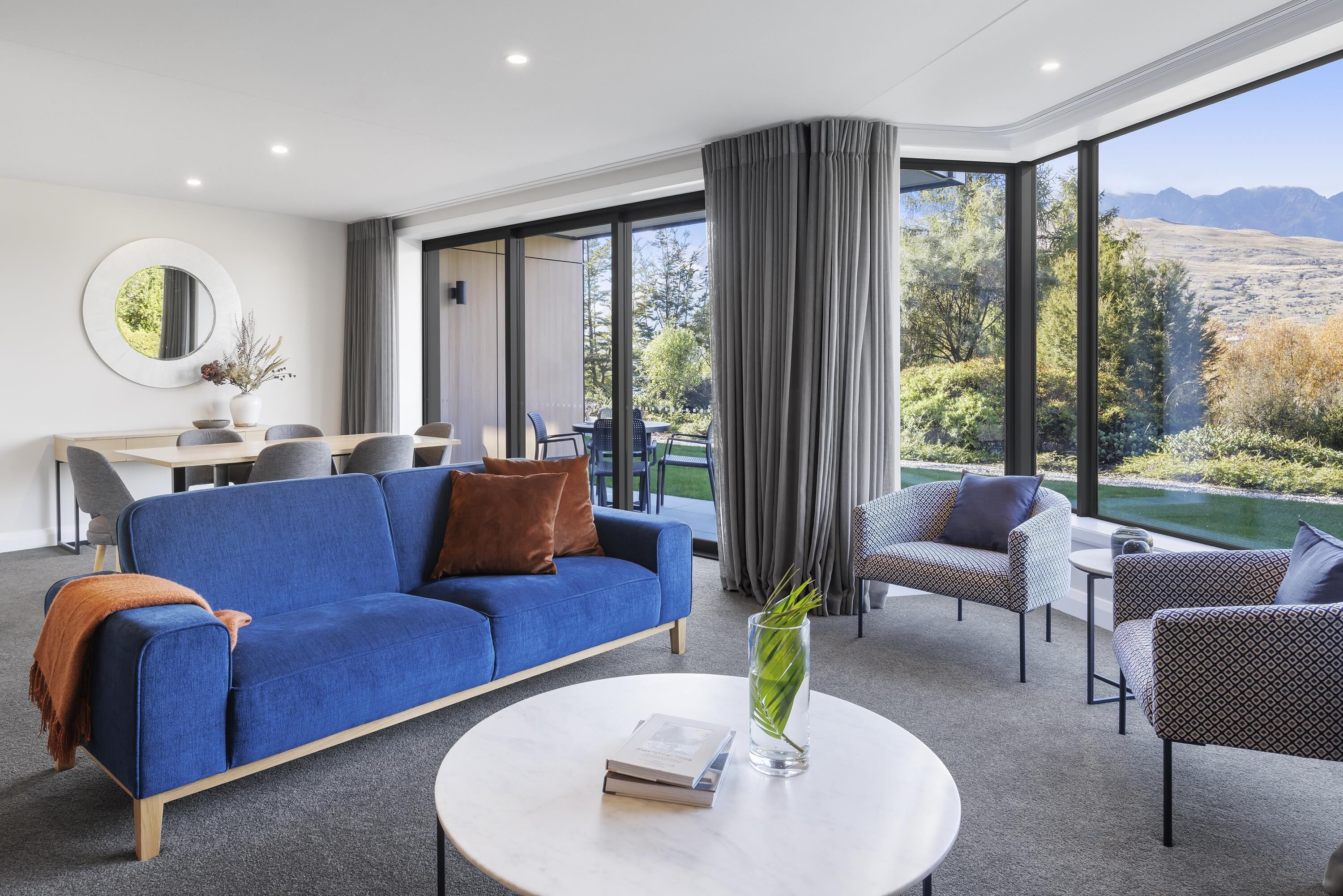 Een moderne woonkamer met een blauwe bank, een ronde salontafel en uitzicht op een bergachtig landschap door grote ramen.