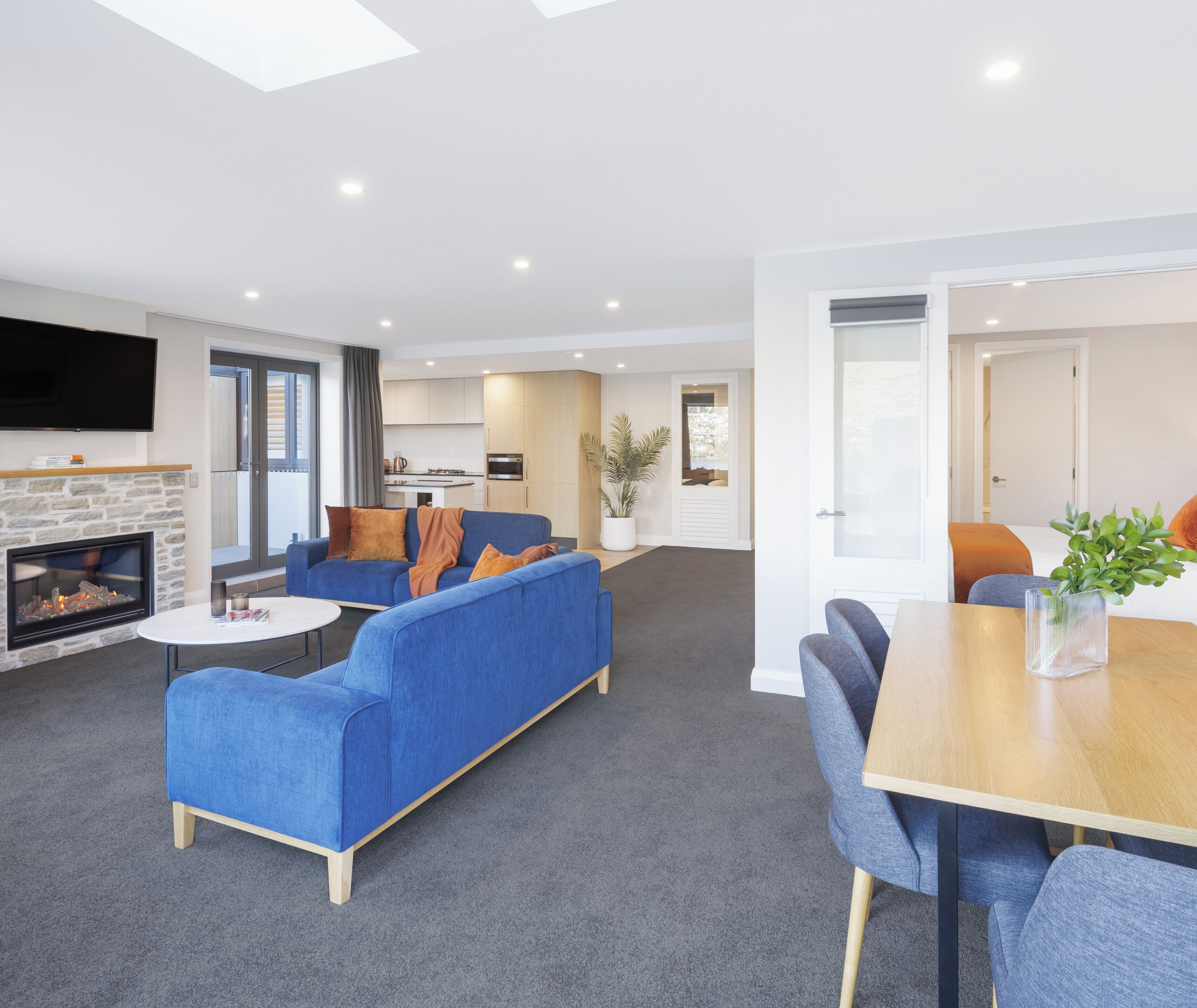 A modern living room with a fireplace, a blue sofa, a dining table with chairs, and a kitchen in the background.