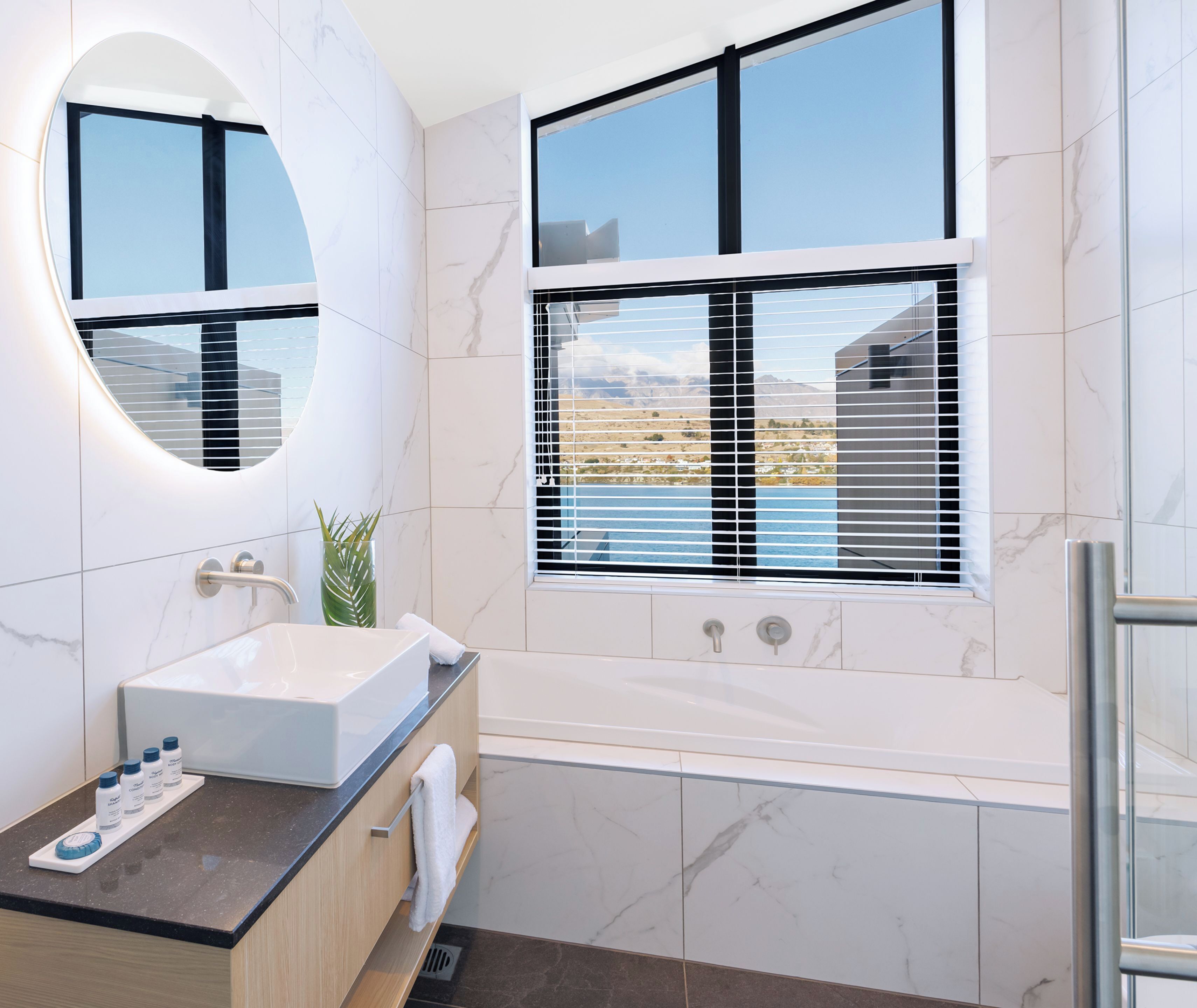 Un baño moderno con un espejo ovalado grande, una bañera independiente y un ventanal con persianas.