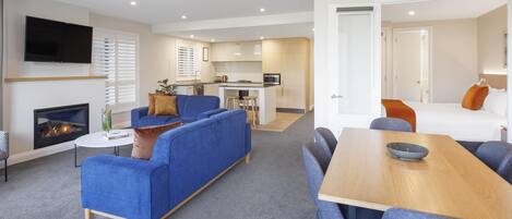 A modern living room with a fireplace, blue sofas, a wooden dining table, and a kitchen in the background.