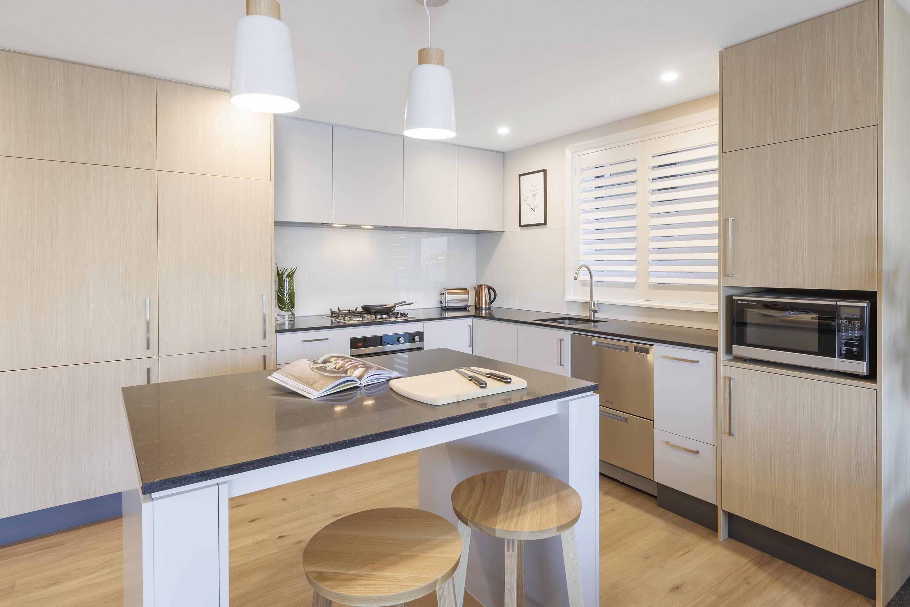 A modern kitchen with light wood cabinets, a central island with stools, a microwave, and a window with blinds.