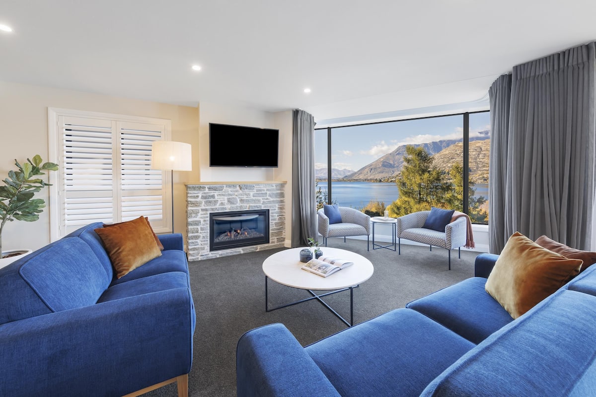 Una sala de estar moderna con chimenea, televisor grande y vistas a un lago y montañas.