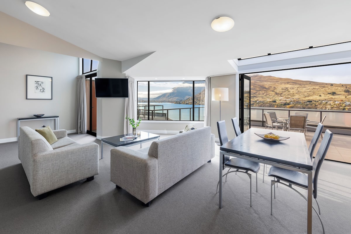 Una sala moderna con sofá, sillón y mesa de comedor. Un ventanal ofrece vistas a un lago y montañas.