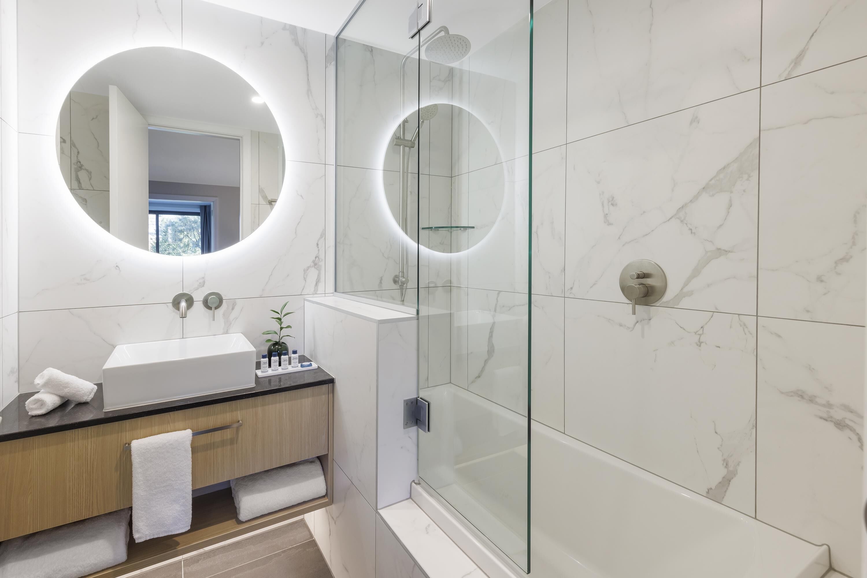 A modern bathroom with a glass-enclosed shower, a round mirror, and a white sink.