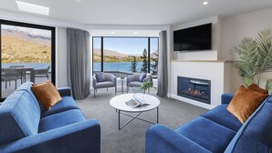 A modern living room with a fireplace, blue sofas, a flat-screen TV, and a large window offering a scenic view of a lake and mountains. - Avani Queenstown Hotel (Queenstown)