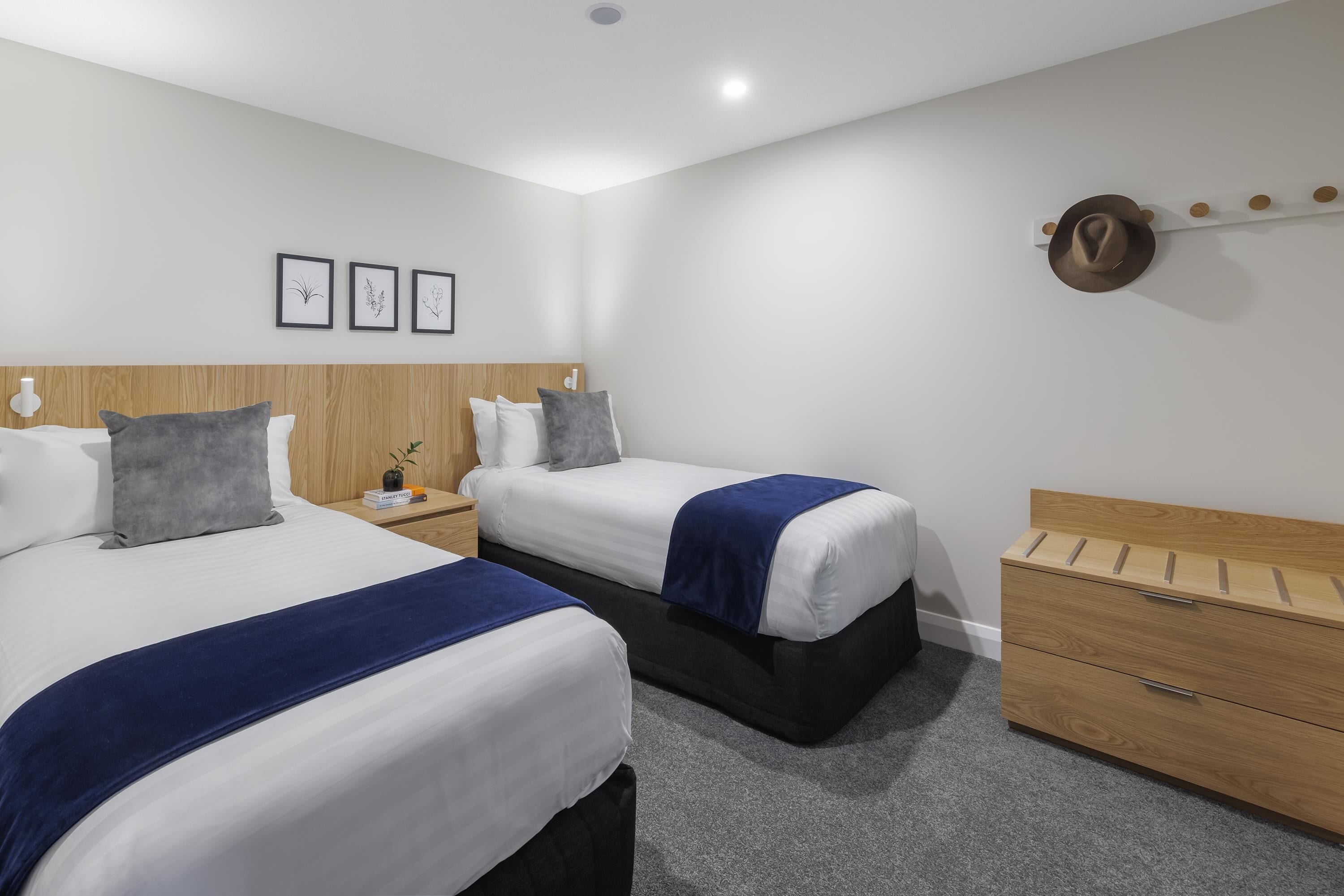 Een hotelkamer met twee bedden, een houten hoofdbord, een nachtkastje met een lamp, een wandkapstok en een kleine plant.
