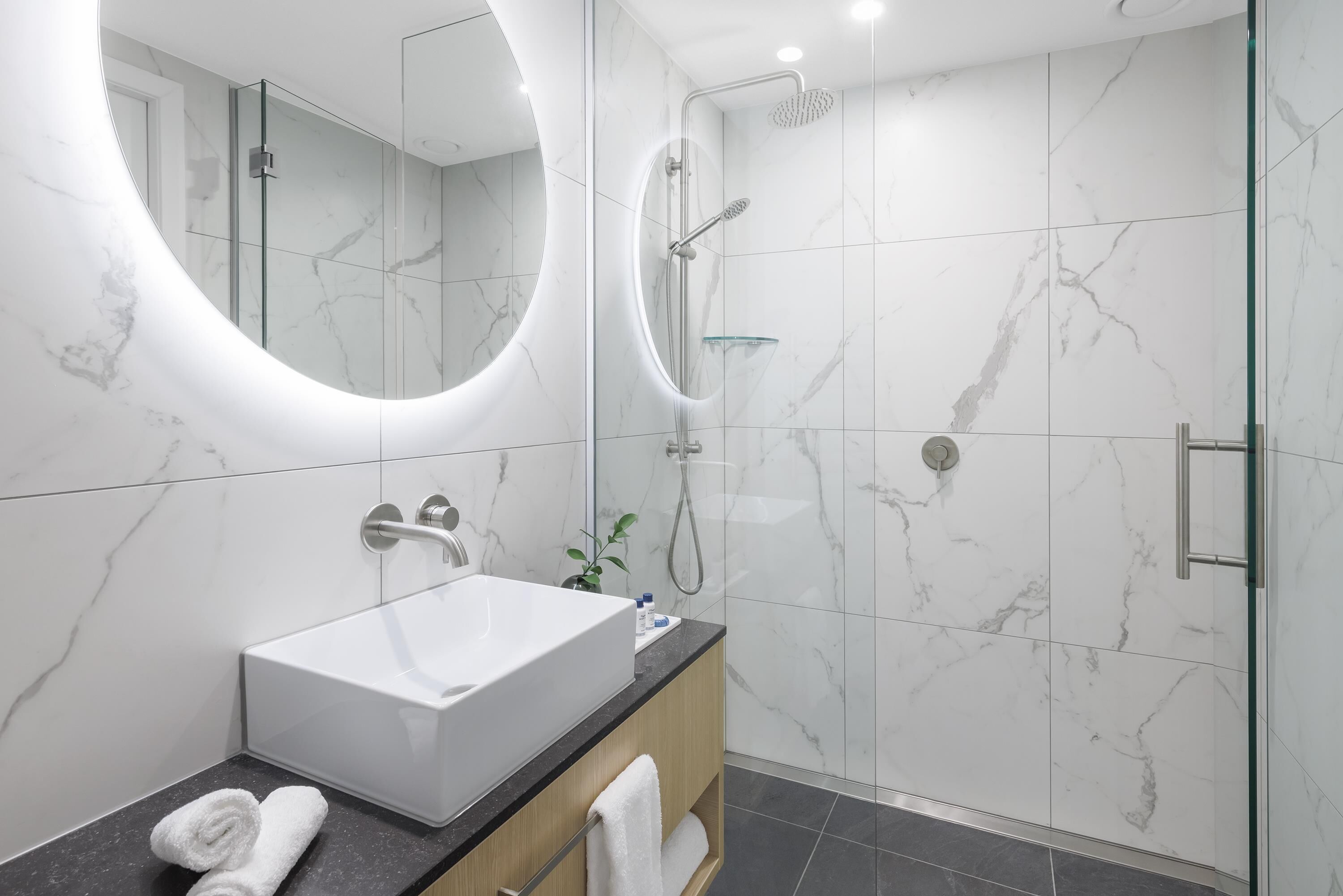 A modern bathroom with a large oval mirror, a white sink, and a glass-enclosed shower.
