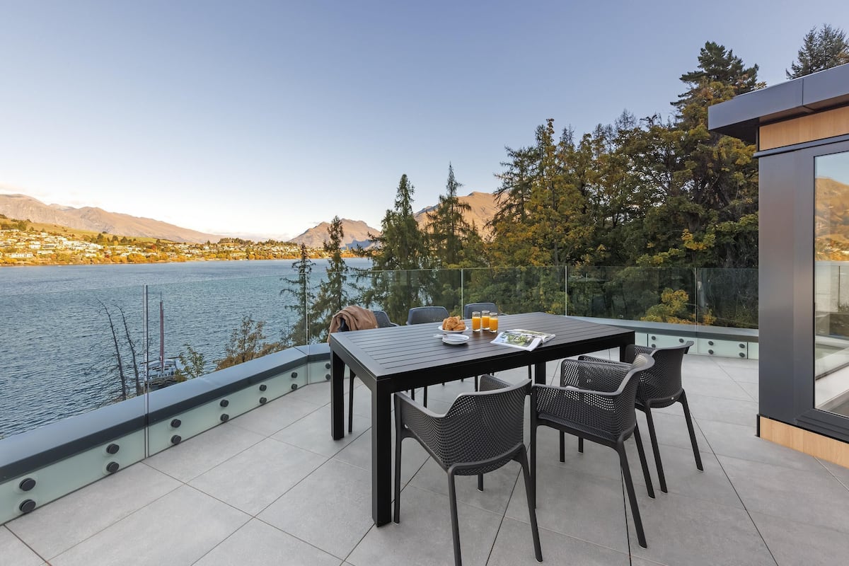 Una terraza con mesa y sillas que da a un lago, rodeada de árboles y montañas.