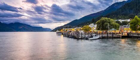 A waterfront dock with boats, a pier, and buildings with lights, surrounded by mountains and a lake.