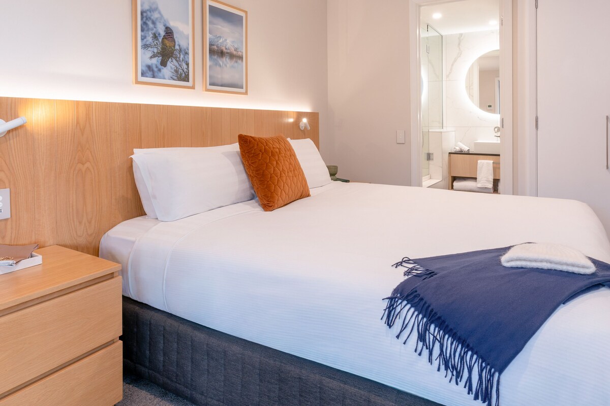 A hotel room with a bed, a nightstand, a bedside table, a mirror, and a wall with two framed pictures.