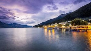 A calm lake with boats docked at a pier, surrounded by mountains and a town with buildings. - Avani Queenstown Hotel (Queenstown)
