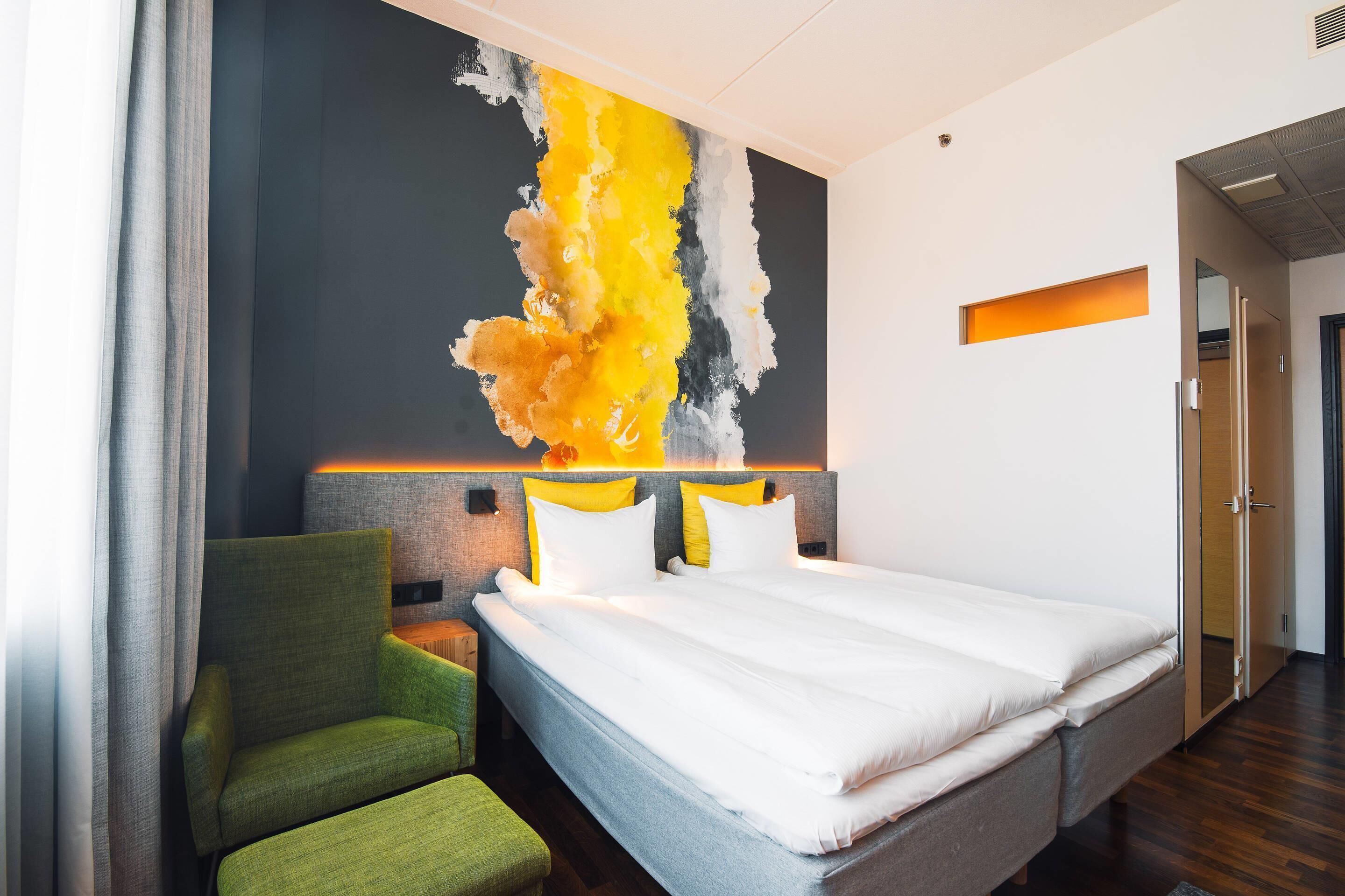 A modern hotel room with a large bed, a green armchair, and a vibrant abstract painting on the wall.
