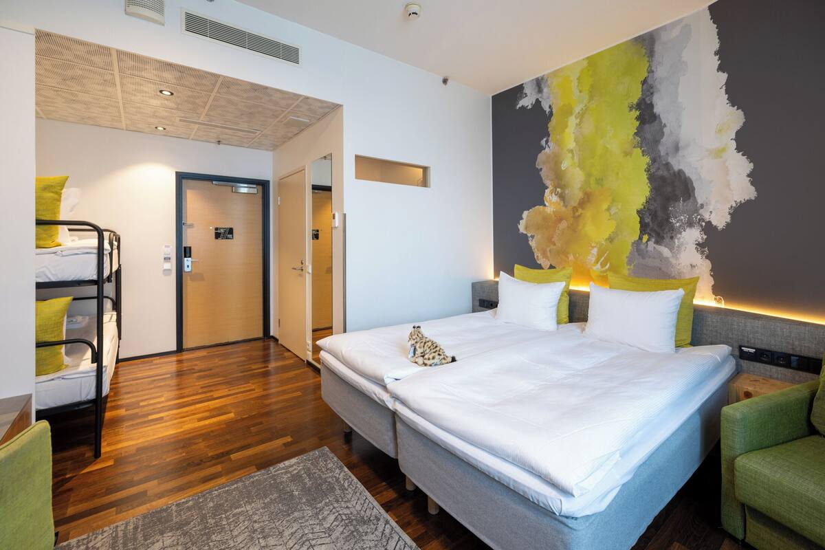 A modern hotel room with a large bed, a bunk bed, a green armchair, and a large abstract painting on the wall.
