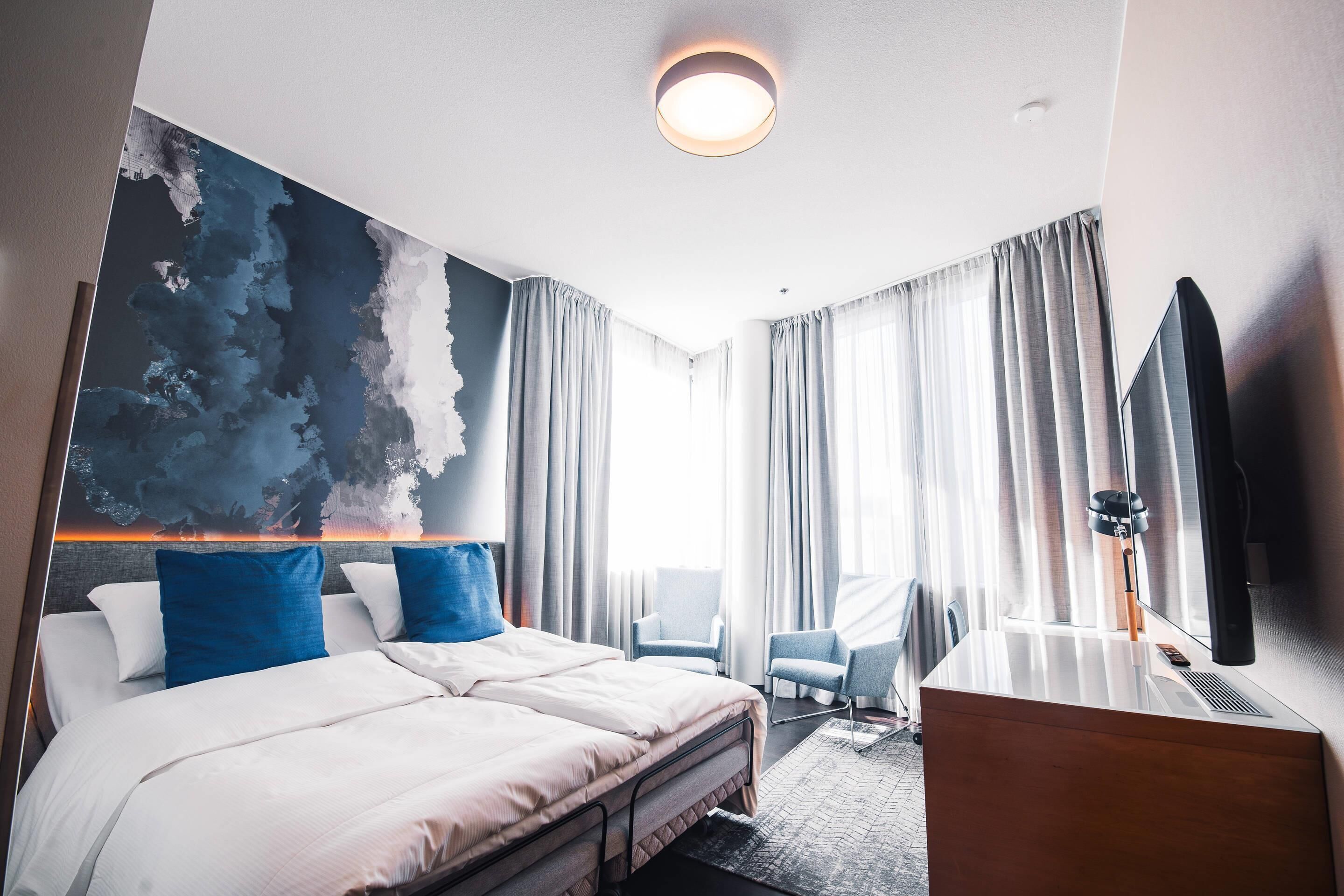 A modern hotel room with a large bed, two chairs, a desk, and a large window with curtains.