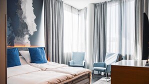 Habitación de hotel con una cama grande, dos sillones, un escritorio y vistas a la ciudad a través de cortinas translúcidas.
