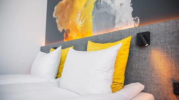 Una cama bien hecha con almohadas blancas y amarillas, un cabecero gris y una pared con un cuadro abstracto de colores.