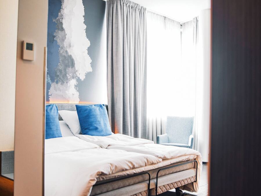 Una habitación de hotel moderna con una cama grande, una silla, una pared con diseño texturizado y una lámpara en el techo.