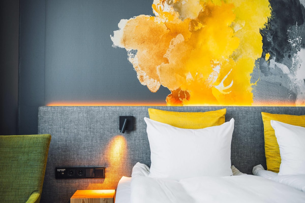 A hotel room with a bed, a nightstand, and a wall-mounted control panel.