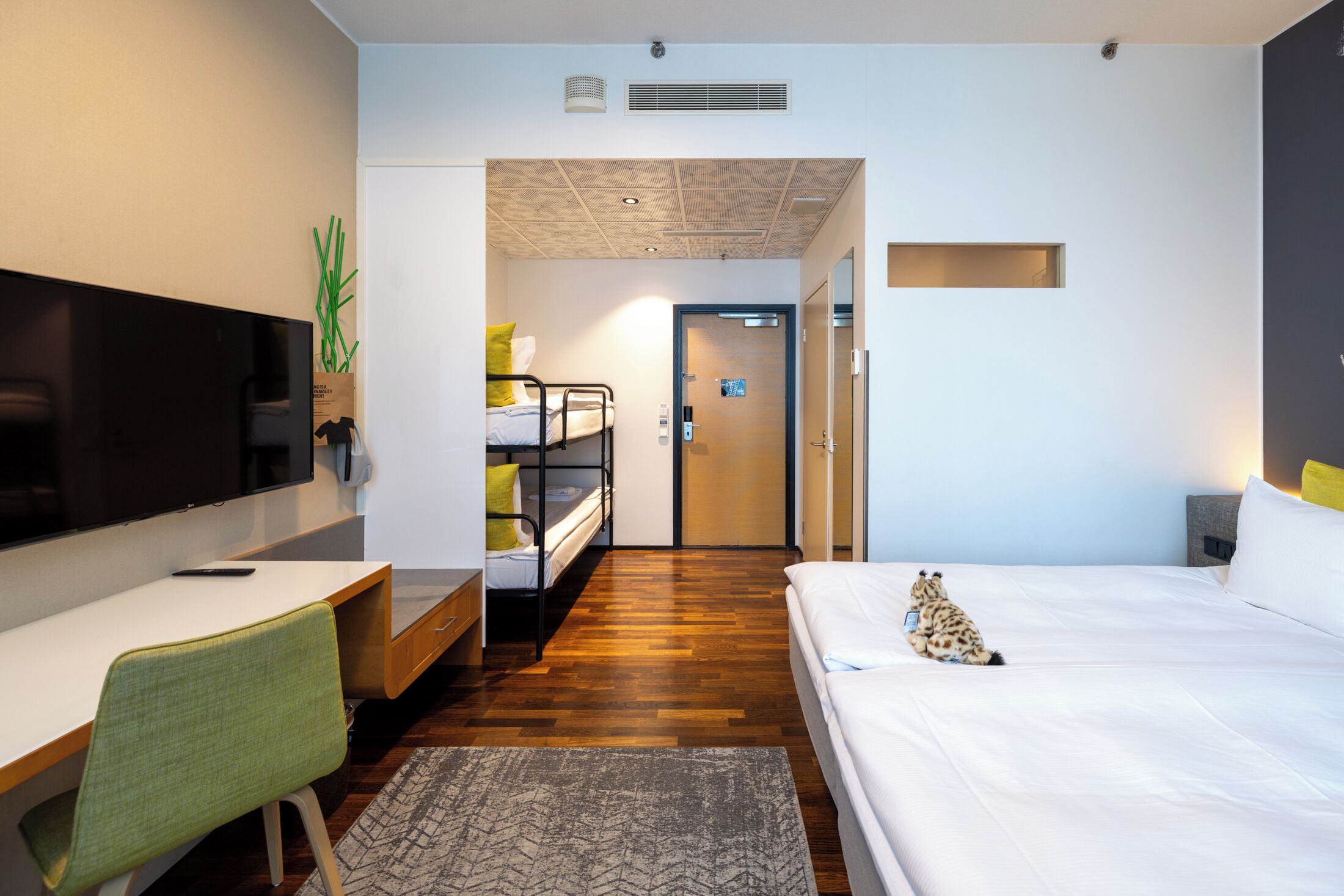 A modern hotel room with a bed, a desk with a chair, a television, and a small plant.