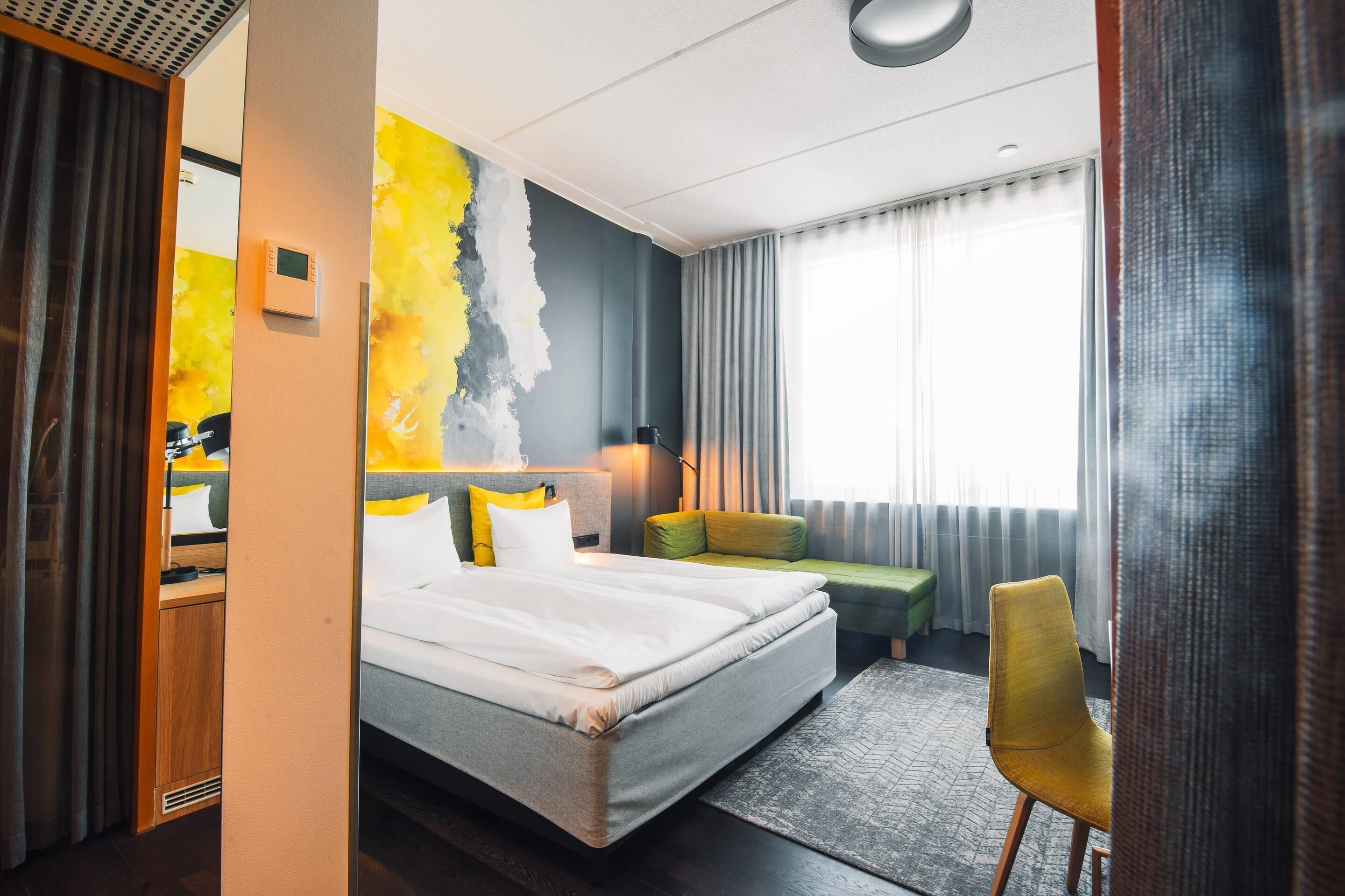 A modern hotel room with a large bed, a green sofa, a yellow chair, and a view of the city.