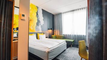 A modern hotel room with a large bed, a green sofa, a yellow chair, and a view of the city.