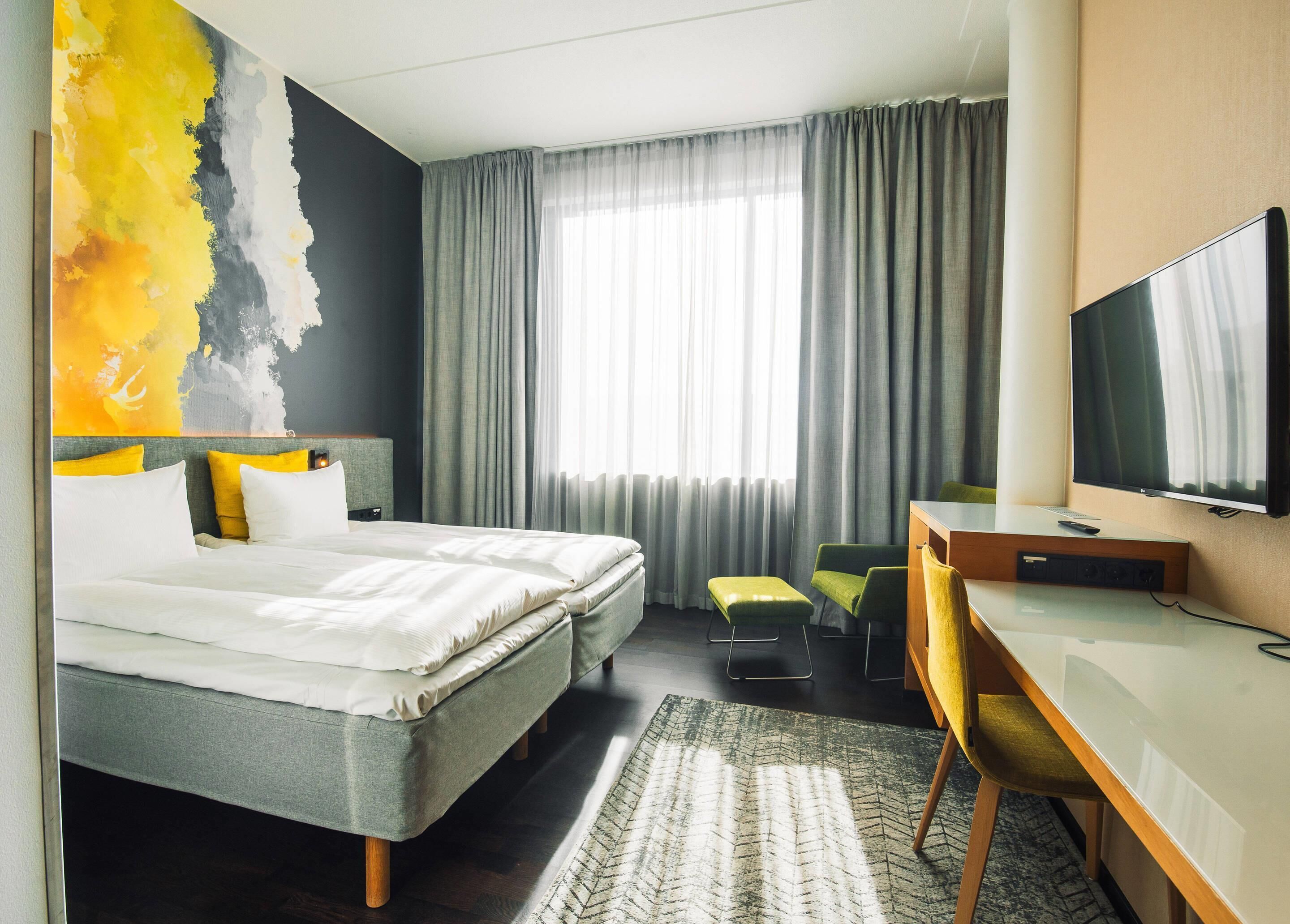 Ein Hotelzimmer mit Bett, Schreibtisch, Fernseher und einem großen Fenster mit Vorhängen.