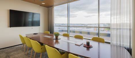 Un salón de conferencias con una gran mesa, sillas amarillas, un televisor de pantalla plana y vistas de la ciudad a través de amplias ventanas.