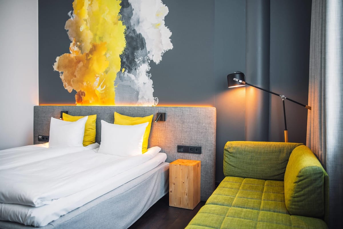 A modern hotel room with a bed, a green sofa, a wooden nightstand, and a wall with a colorful abstract painting.