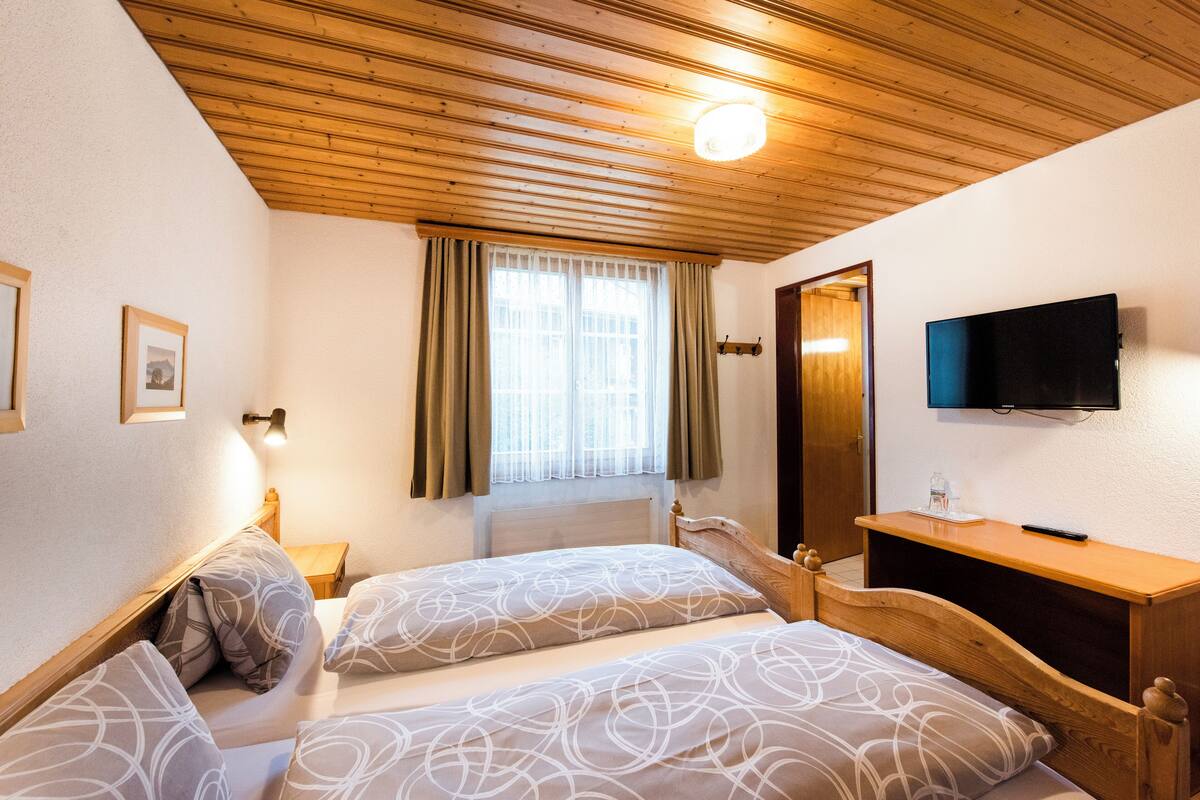 Una camera da letto con due letti, soffitto in legno, una finestra con tende, un comodino e una TV a parete, oltre a un altro mobiletto in legno.