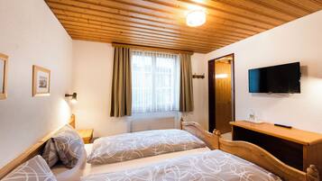 A bedroom with two beds, a wooden ceiling, a window with curtains, a bedside table, a TV mounted on the wall, and a wooden nightstand.